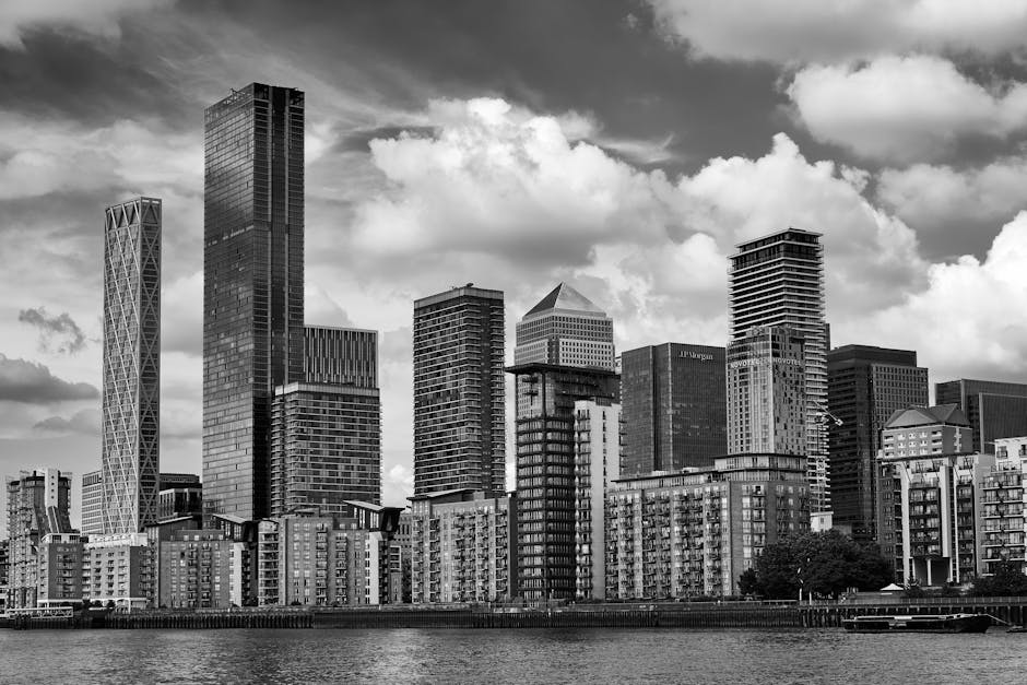 A black and white view of a modern city skyline featuring a variety of high-rise buildings with different architectural styles, including rectangular glass towers and a tower with a crisscrossed facade, situated along a waterfront. Below the skyline, there are mid-rise residential buildings with numerous balconies and windows, built with brick or concrete finishes. The scene is set under a partly cloudy sky with large, fluffy clouds scattered across, and the water in the foreground reflects some of the buildings. The overall composition emphasizes the contrast between the towering structures and the lower residential blocks, illustrating urban development typical of a commercial and residential area. This environment exemplifies the kind of cityscape that can benefit from efficient private waste disposal or rubbish removal services like those offered by Waste Removal Canary Wharf, especially in areas with high-density buildings.