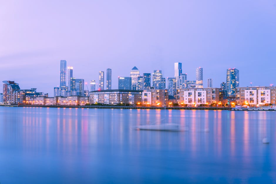 A panoramic view of a modern city skyline at dusk, featuring high-rise office buildings and residential apartments with glass facades illuminated by exterior lighting. The buildings are situated along the water's edge, with their reflections visible on the calm surface of a river or harbor in the foreground. Some structures have distinctive architectural features, such as a pyramid-shaped roof and varied heights, set against a soft pastel sky transitioning from light purple to pale blue. In the lower part of the image, there are low-rise buildings with white and beige exteriors and numerous evenly spaced orange streetlights along the waterfront, creating a warm glow that contrasts with the cooler tones of the buildings and water. The scene provides an urban environment where private waste removal services like Waste Removal Canary Wharf might be involved in maintaining cleanliness, especially in areas adjacent to commercial and residential properties near the water, emphasizing the importance of efficient rubbish collection in cityscapes.