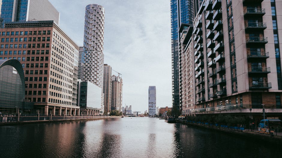 A black and white view of a modern city skyline featuring a variety of high-rise buildings with different architectural styles, including rectangular glass towers and a tower with a crisscrossed facade, situated along a waterfront. Below the skyline, there are mid-rise residential buildings with numerous balconies and windows, built with brick or concrete finishes. The scene is set under a partly cloudy sky with large, fluffy clouds scattered across, and the water in the foreground reflects some of the buildings. The overall composition emphasizes the contrast between the towering structures and the lower residential blocks, illustrating urban development typical of a commercial and residential area. This environment exemplifies the kind of cityscape that can benefit from efficient private waste disposal or rubbish removal services like those offered by Waste Removal Canary Wharf, especially in areas with high-density buildings.