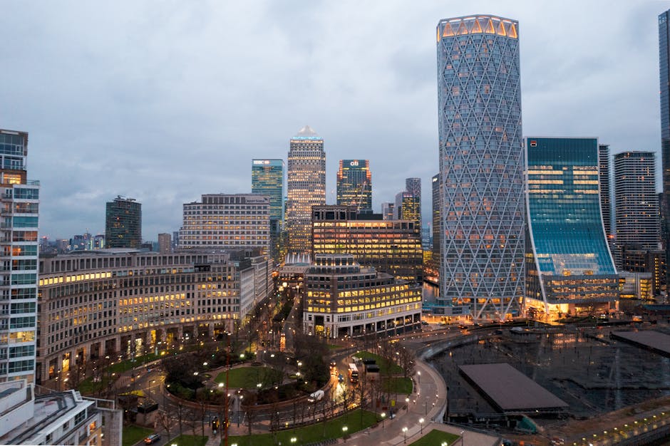 A cityscape featuring modern skyscrapers and office buildings in a business district during dusk, with the tallest structure on the right displaying a distinctive geometric lattice pattern on its glass facade. Surrounding buildings vary in height and architectural style, with some featuring illuminated windows and reflective glass surfaces. The streets below are illuminated by streetlights, and a small park with pathways, benches, and trees is visible in the foreground, indicating an urban environment that may be serviced by independent waste removal companies like Waste Removal Canary Wharf. The overall scene is characterized by a cloudy sky, subdued natural light, and the glow of office lighting reflecting off the buildings, creating an atmospheric view of modern urban development suitable for articles related to city waste management or private rubbish collection services.