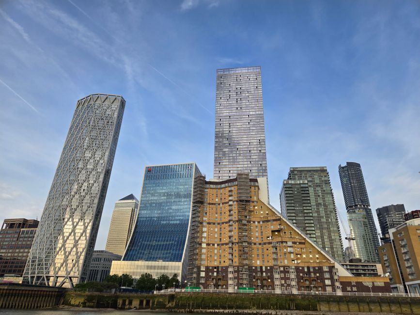 A wide-angle view of several high-rise office buildings in Canary Wharf, London, with a prominent modern skyscraper featuring a glass facade and grid-like pattern at the center. To the left, a distinctive building with a diagonal cross-braced steel exterior and a curved shape. Other surrounding structures include a blue-tinted glass tower, a beige stone building, and additional tall office blocks with reflective windows. The scene is captured during daytime with clear skies, showing a mix of natural light and slight reflections on the glass surfaces. The environment suggests an urban commercial district with minimal ground-level details visible, emphasizing the height and architectural diversity of the skyline. This setting relates to private waste management and on-site clearance services that companies like Waste Removal Canary Wharf provide for such corporate environments, ensuring efficient rubbish collection within busy cityscapes.