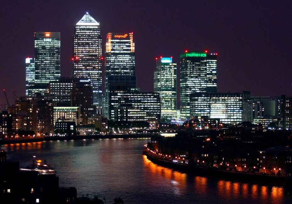 A nighttime cityscape of Canary Wharf, featuring illuminated modern skyscrapers with glass facades reflecting city lights, including the prominent Citigroup and HSBC buildings, situated along the River Thames. The water’s surface shows reflections of the bright lights from the buildings and surrounding streetlights, with a dark sky overhead. In the foreground, there are low-rise buildings and a docked boat or barge with orange and yellow lights along the shoreline. The scene captures the urban environment typical of a financial district with a mix of corporate high-rises and waterfront infrastructure, relevant to private waste handling or outside agency rubbish removal services such as Waste Removal Canary Wharf operating in this commercial area.