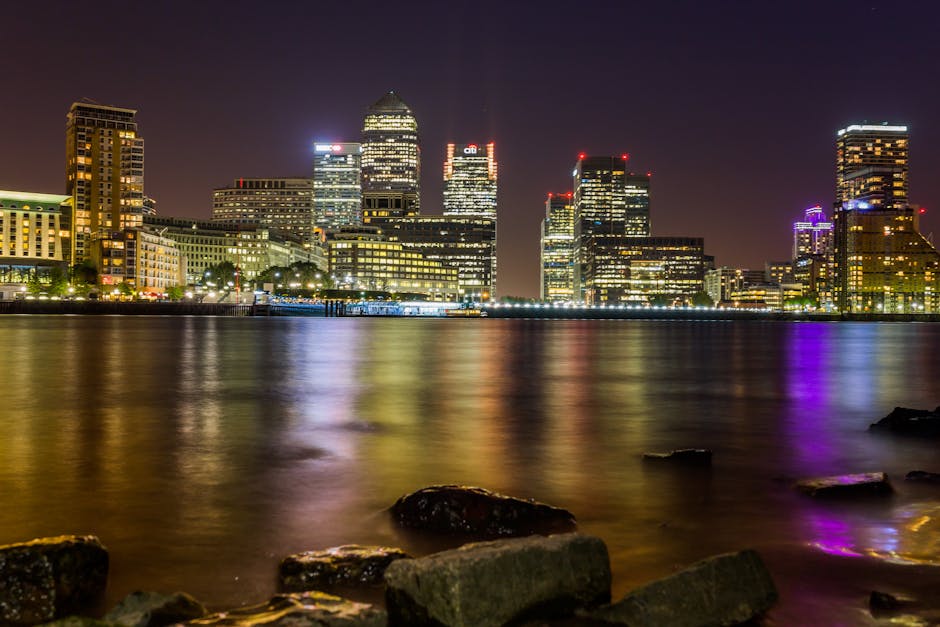 A nighttime cityscape of Canary Wharf, featuring illuminated modern skyscrapers with glass facades reflecting city lights, including the prominent Citigroup and HSBC buildings, situated along the River Thames. The water’s surface shows reflections of the bright lights from the buildings and surrounding streetlights, with a dark sky overhead. In the foreground, there are low-rise buildings and a docked boat or barge with orange and yellow lights along the shoreline. The scene captures the urban environment typical of a financial district with a mix of corporate high-rises and waterfront infrastructure, relevant to private waste handling or outside agency rubbish removal services such as Waste Removal Canary Wharf operating in this commercial area.