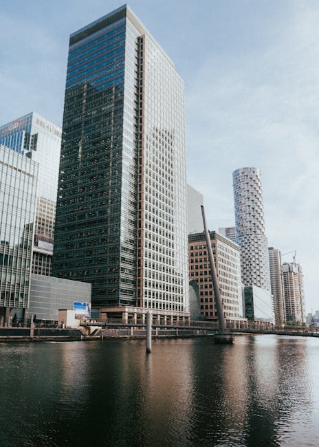 The image depicts a cluster of modern high-rise buildings situated alongside a waterway, with the tallest structure predominantly composed of dark glass windows reflecting the sky. To the right, a distinct white skyscraper features a rounded, textured facade with a pattern of small, square windows creating a grid-like appearance. Surrounding buildings vary in height and design, including some with light-colored exteriors and grid-like window patterns. The scene is illuminated by natural daylight, with a clear sky that has soft, light clouds, enhancing the reflective quality of the glass facades. In the foreground, the calm water reflects the buildings, with a narrow strip of pavement and metal railing visible along the water's edge. The setting appears to be an urban business district, characteristic of Canary Wharf, and contextualised by the presence of such towering structures. This cityscape underscores the importance of efficient rubbish management in dense commercial areas, where regular waste collection and on-site clearance services, such as those provided by Waste Removal Canary Wharf, are vital for maintaining cleanliness and order in a busy metropolitan environment.