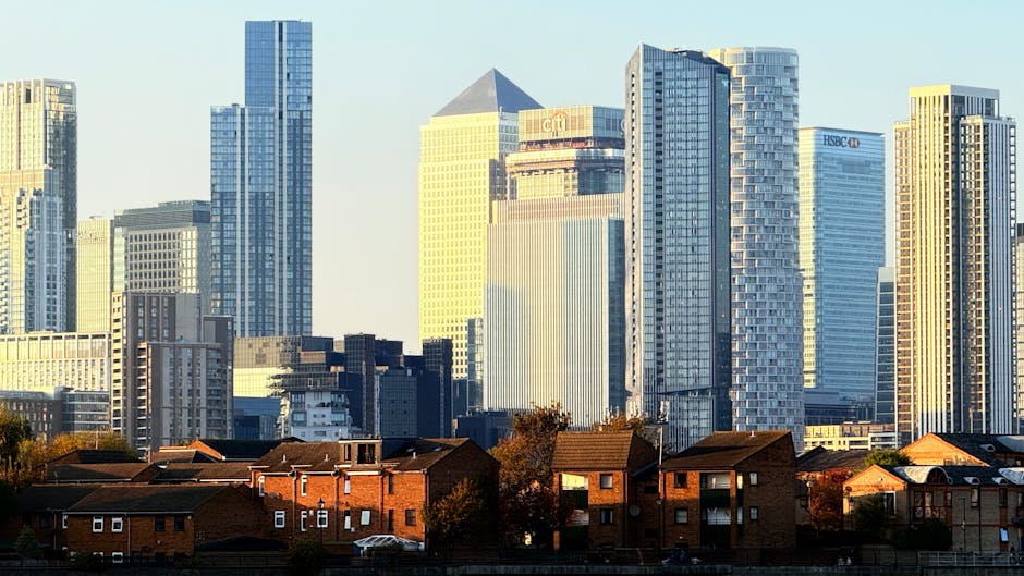 A cityscape featuring a foreground of low-rise red-brick residential buildings with pitched roofs, windows, and small trees alongside. Behind these, a cluster of modern high-rise office towers and skyscrapers made of glass, steel, and concrete reach upward, reflecting daylight with their sleek finishes and large window panels. The taller buildings display various architectural styles, including rectangular and curved facades, with some bearing visible signage such as HSBC and Citi. The scene is illuminated by natural sunlight, casting subtle shadows and highlighting the textures of both the older houses and the contemporary city skyline. The background sky is clear, and the overall environment suggests an urban area where private waste collection services, like Waste Removal Canary Wharf, may assist in managing rubbish and bulky waste from residential and commercial properties, complementing or offering alternative waste handling solutions to municipal rubbish collection.