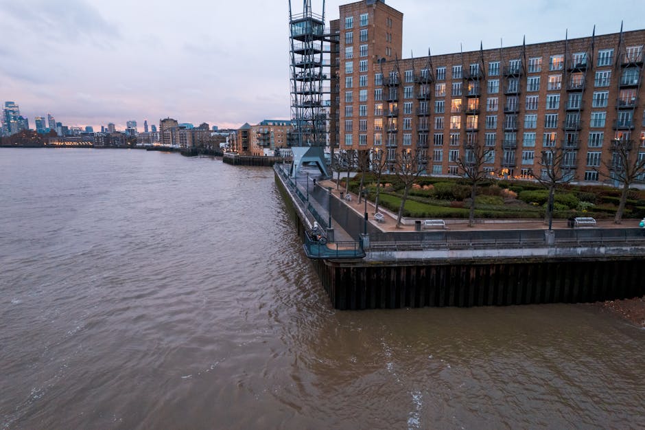 The image depicts a cluster of modern high-rise buildings situated alongside a waterway, with the tallest structure predominantly composed of dark glass windows reflecting the sky. To the right, a distinct white skyscraper features a rounded, textured facade with a pattern of small, square windows creating a grid-like appearance. Surrounding buildings vary in height and design, including some with light-colored exteriors and grid-like window patterns. The scene is illuminated by natural daylight, with a clear sky that has soft, light clouds, enhancing the reflective quality of the glass facades. In the foreground, the calm water reflects the buildings, with a narrow strip of pavement and metal railing visible along the water's edge. The setting appears to be an urban business district, characteristic of Canary Wharf, and contextualised by the presence of such towering structures. This cityscape underscores the importance of efficient rubbish management in dense commercial areas, where regular waste collection and on-site clearance services, such as those provided by Waste Removal Canary Wharf, are vital for maintaining cleanliness and order in a busy metropolitan environment.